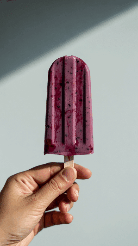 A hand holding a single, creamy Blueberry, Thyme and Sweet Cream Ice Pop.