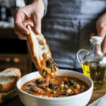 A person dipping crusty bread into a warm bowl of Sausage, Beans and Broccoli Rabe Soup.