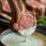 A hand dipping a cooked lamb chop into a bowl of creamy yogurt sauce.
