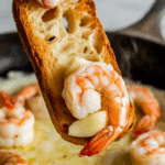 A hand dipping crusty bread into the garlic-butter sauce from the roasted shrimp.