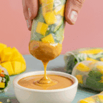 A hand dipping a Mango Summer Roll into a bowl of peanut sauce.