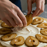 Hands carefully arranging roasted pear halves in a circular pattern on top of a layer of almond cream on a raw pastry dough.