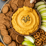 A beautiful platter with a bowl of Pumpkin Dip in the center, surrounded by a variety of cookies, fruits, and pretzels for dipping at a party.