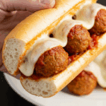 A hand holding a large, delicious, and messy meatball sub sandwich made with homemade turkey meatballs and melted cheese.