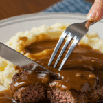 A fork cutting into a tender slice of Salisbury Steak with mushroom gravy and mashed potatoes.