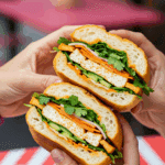 A hand holding a large, delicious, and overflowing homemade Tofu Banh Mi sandwich, about to take a bite.