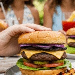 A hand holding a large, delicious, and fully assembled homemade Portobello Mushroom Burger at a summer barbecue.