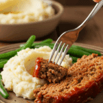 A beautiful plate of a homemade, thick, and saucy slice of BBQ Meatloaf, served with creamy mashed potatoes and green beans for a family dinner.
