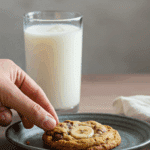 A hand reaching to take a warm, homemade Banana Chocolate Chip Cookie from a plate that is next to a tall, cold glass of milk.