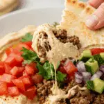A piece of a torn pita bread being used to scoop up a delicious bite of a homemade Ground Beef Hummus Bowl at a casual family dinner.