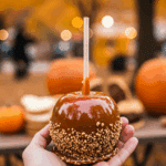 A hand holding a single, beautiful, and glossy, homemade Caramel Apple at a fun and festive outdoor fall festival.