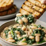 A cozy bowl of homemade, creamy White Beans and Greens being enjoyed with a large piece of crusty sourdough bread for dipping.