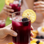 A hand holding an elegant glass of a homemade, purple, and refreshing Blackberry Lemonade Punch at a fun and festive outdoor party.